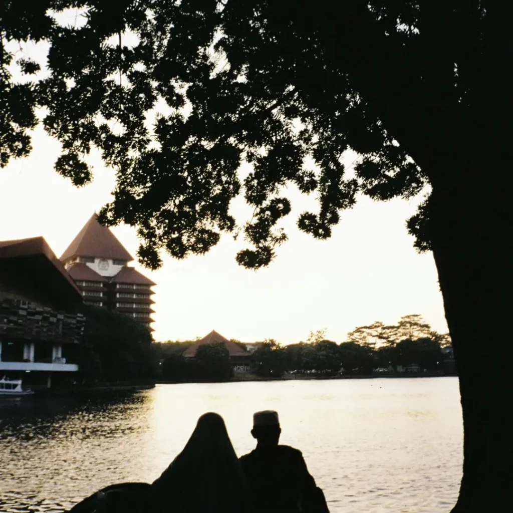 Dua orang sedang duduk di depan danau UI dan memandang salah satu gedung Universitas Indonesia dalam artikel Landmark di Kota Depok di website ecotown.id