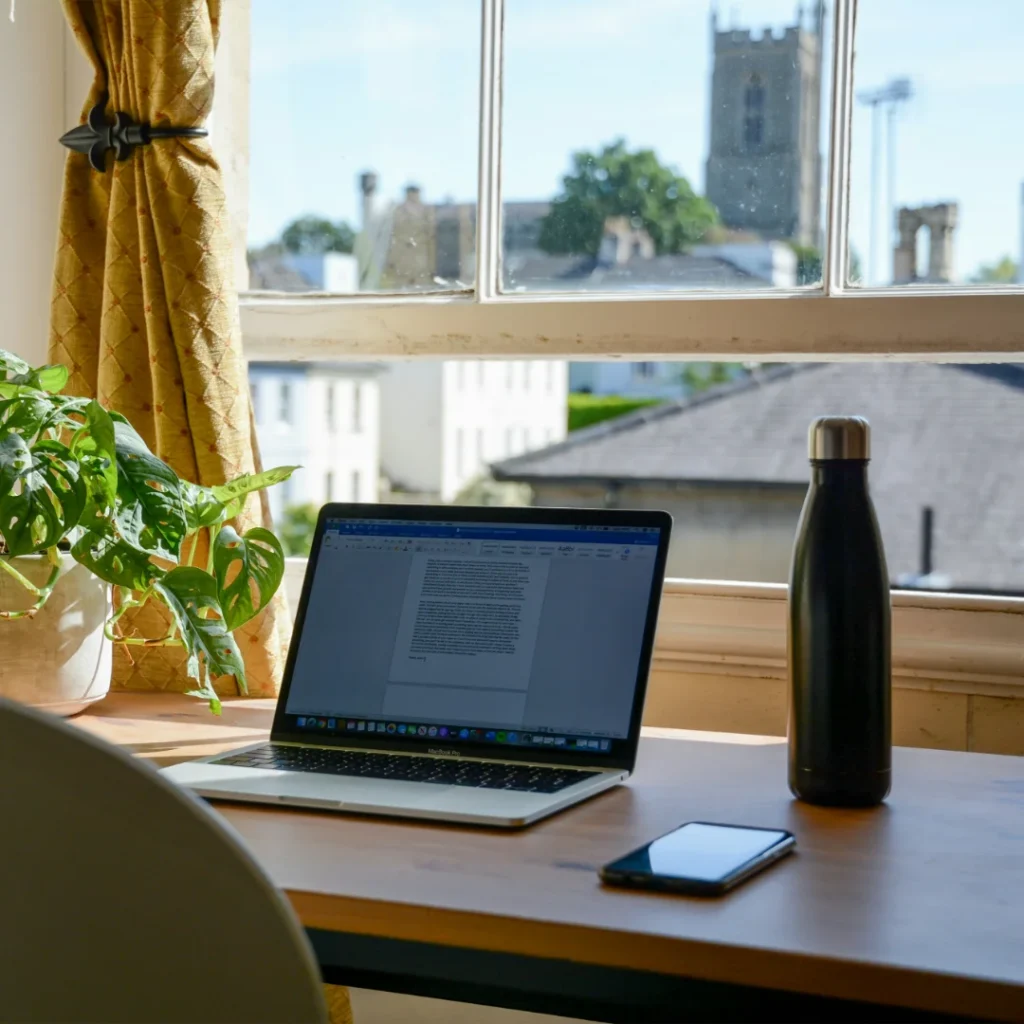 Laptop, handphone, dan tumbler di atas sebuah meja yang menghadap ke luar rumah dalam artikel Rumah yang Cocok Untuk Work From Home di website Ecotown.id