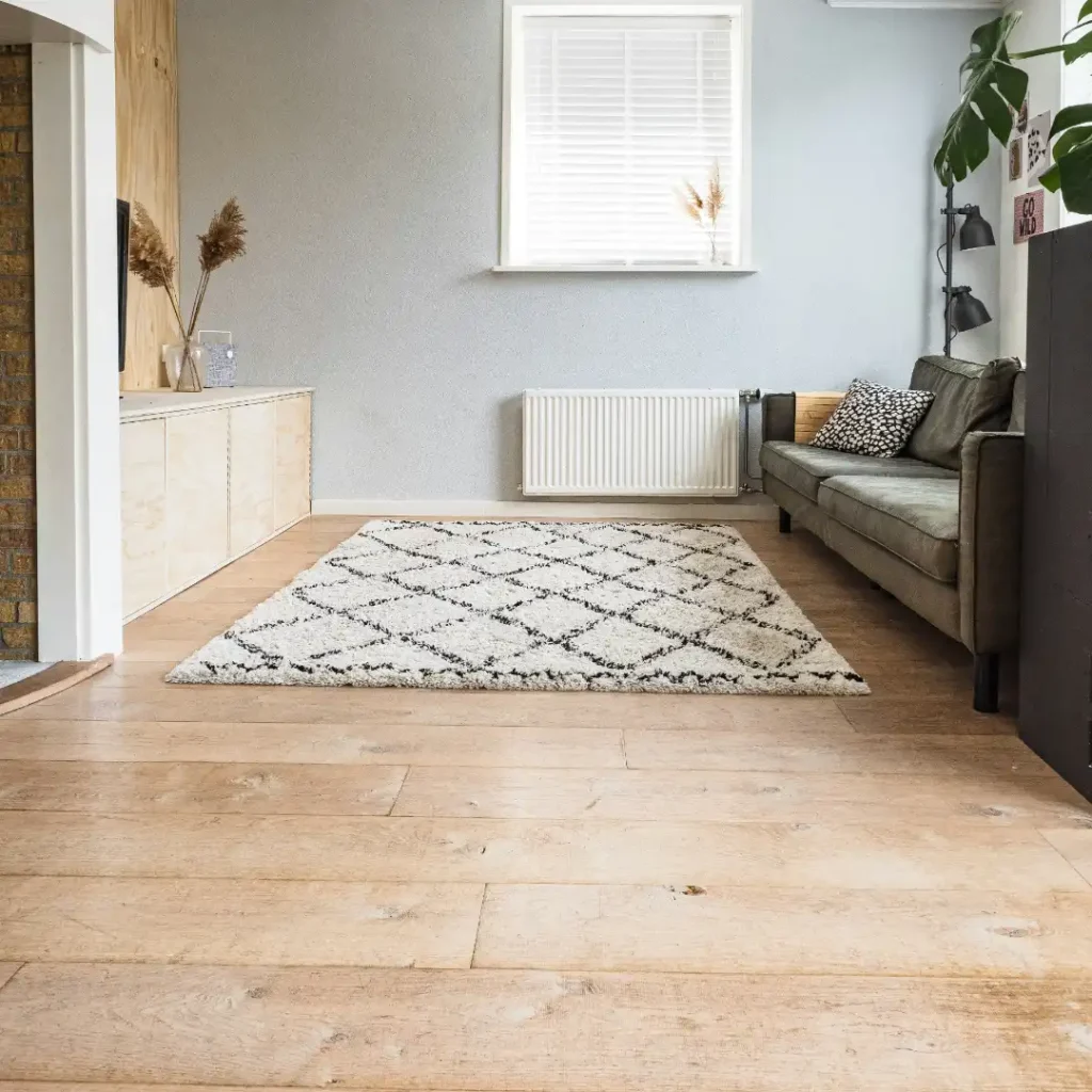 Ruang keluarga minimalis dengan lantai kayu hangat, karpet bermotif geometris, serta sofa abu-abu dalam artikel "Jenis Lantai Rumah" di website Ecotown.id