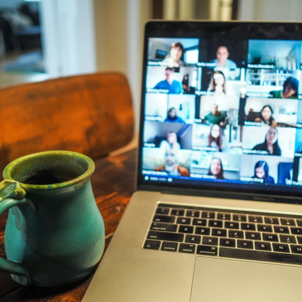 Sebuah laptop yang menampilkan layar panggilan konferensi video dengan banyak peserta, diletakkan di atas meja kayu berdampingan dengan cangkir keramik berwarna hijau. Gambar ini berada dalam artikel "Produktivitas Kerja Dari Rumah" di website Ecotown.id