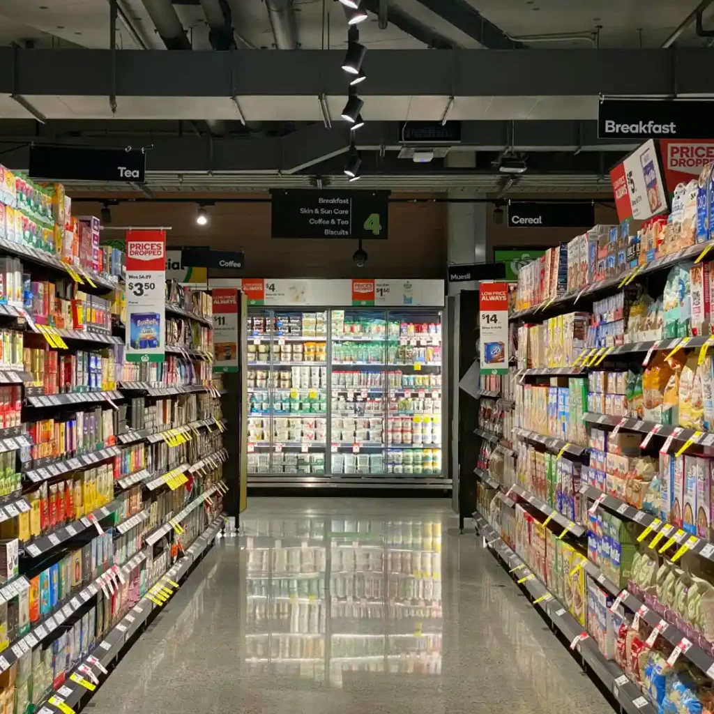 Lorong supermarket yang terang dan bersih dengan rak-rak di kedua sisi yang memajang berbagai produk makanan kemasan seperti sereal dan teh, mengarah pada deretan lemari pendingin di ujung lorong. Gambar ini berada dalam artikel "Supermarket di Sawangan Depok" di website Ecotown.id
