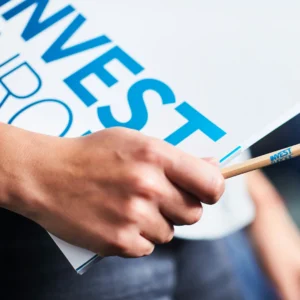 Bidikan jarak dekat tangan seseorang yang sedang memegang ujung dokumen atau papan putih bertuliskan "INVEST" berwarna biru muda bersama dengan sebuah pensil kayu. Gambar ini berada dalam artikel "Investasi Ruko vs Rumah Hunian" di website Ecotown.id
