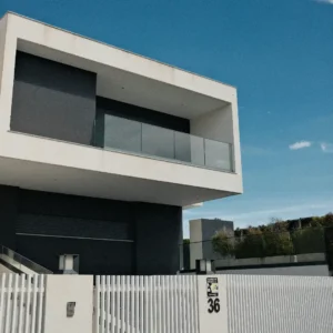 Fasad rumah modern minimalis bertingkat dua dengan kombinasi warna putih dan abu-abu gelap, balkon berdinding kaca, serta pagar putih. Gambar ini berada dalam artikel "Jenis Atap Rumah Minimalis" di website Ecotown.id