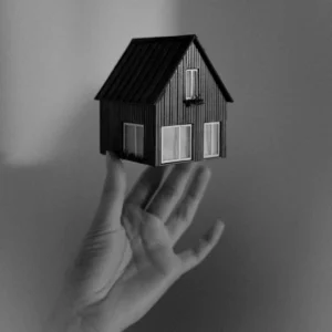 Sebuah foto bernuansa hitam putih yang menampilkan ujung jari dari satu tangan yang terbuka ke atas untuk menopang sebuah miniatur rumah beratap miring. Gambar ini berada dalam artikel "Millenial dan Gen-Z Beli Rumah" di website Ecotown.id