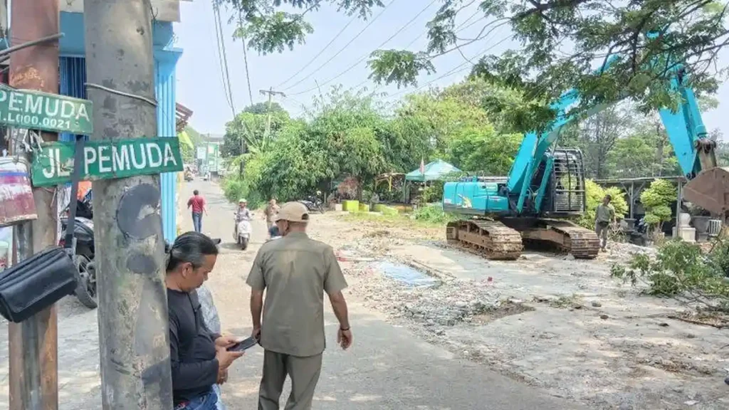 Suasana pengerjaan atau pelebaran jalan di mana sebuah ekskavator biru toska sedang mengeruk tanah di sisi jalan, sementara beberapa warga terlihat berjalan dan berdiri memperhatikan di dekat tiang listrik beton yang memiliki plang hijau bertuliskan "JL PEMUDA". Gambar ini berada dalam artikel "Pelebaran Jalan Enggram-Pemuda Sawangan" di website Ecotown.id