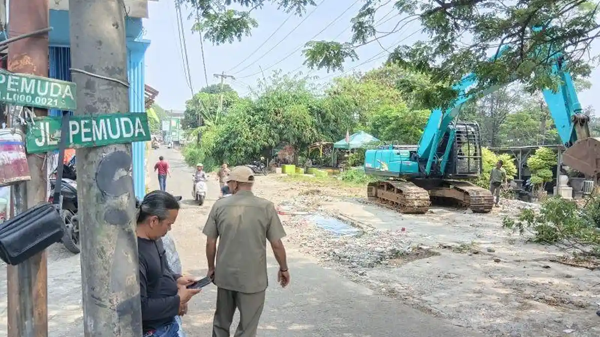 Suasana pengerjaan atau pelebaran jalan di mana sebuah ekskavator biru toska sedang mengeruk tanah di sisi jalan, sementara beberapa warga terlihat berjalan dan berdiri memperhatikan di dekat tiang listrik beton yang memiliki plang hijau bertuliskan "JL PEMUDA". Gambar ini berada dalam artikel "Pelebaran Jalan Enggram-Pemuda Sawangan" di website Ecotown.id