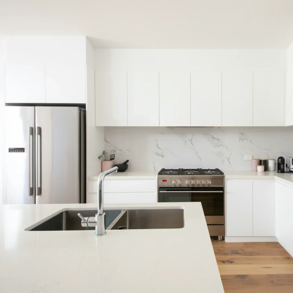 Ruangan dapur berdesain minimalis dan modern yang sangat terang, dilengkapi dengan lemari putih tanpa pegangan, meja dapur putih bersih, dinding pelindung cipratan air (backsplash) bermotif marmer, serta perabotan baja tahan karat seperti kulkas dan kompor oven. Gambar ini berada dalam artikel "Renovasi Dapur Minimalis" di website Ecotown.id