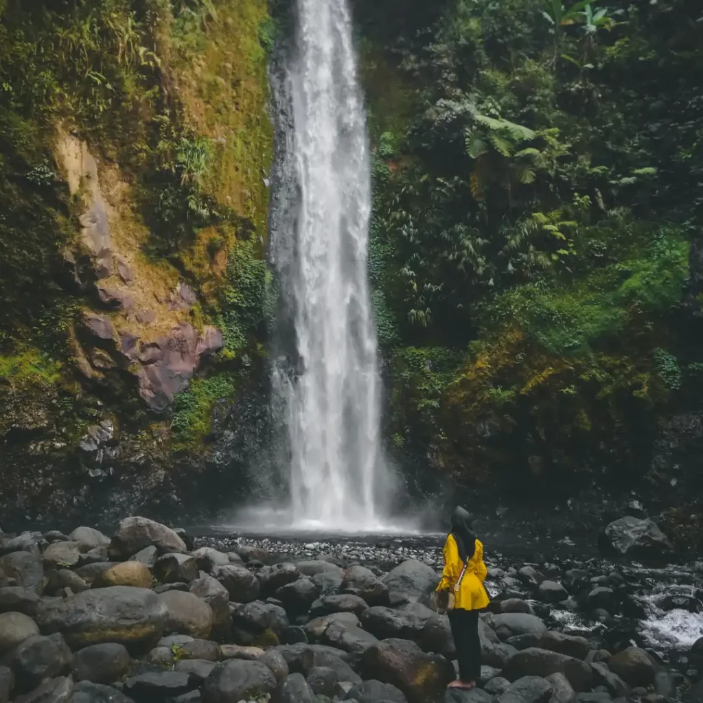 Seorang wanita berhijab dengan baju kuning berdiri di antara bebatuan di bawah air terjun tinggi yang dikelilingi pepohonan hijau rindang. Gambar ini berada dalam artikel "Tempat Wisata di Depok Selatan" di website Ecotown.id