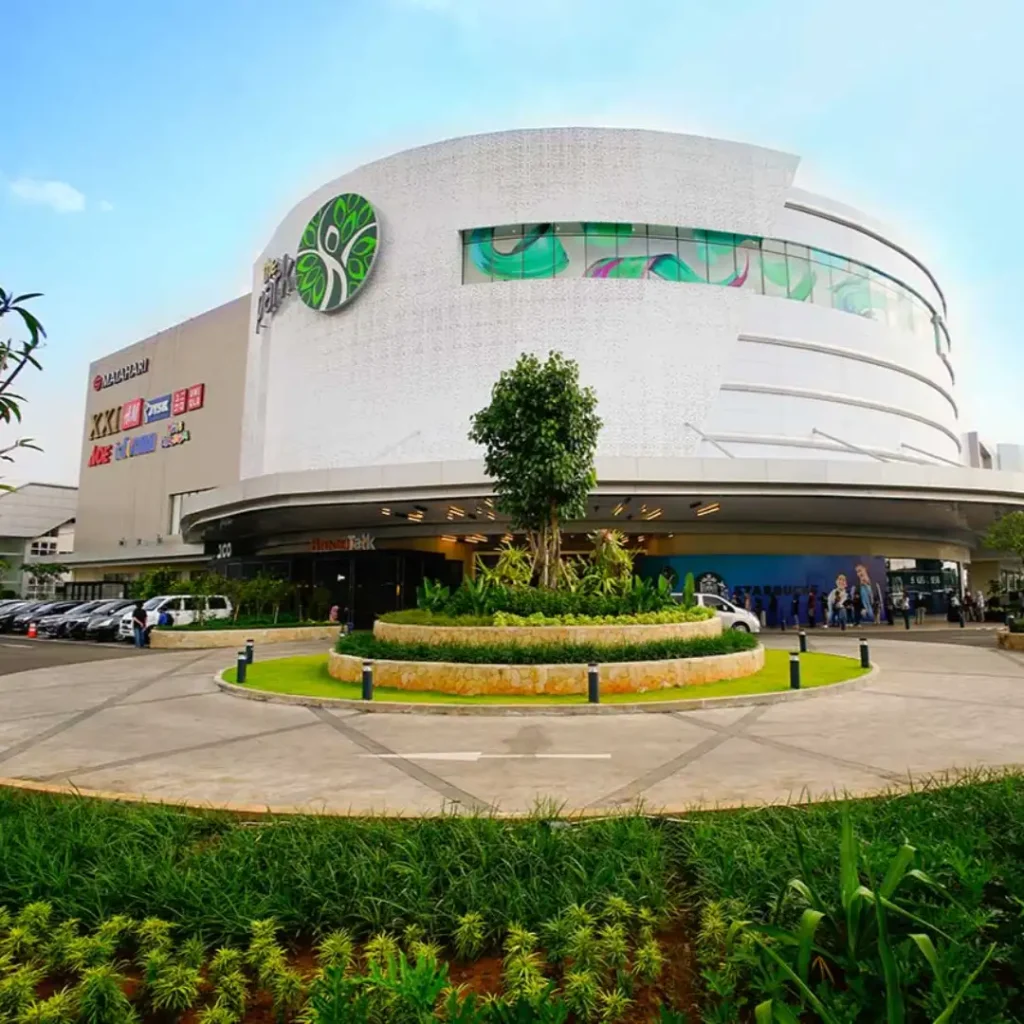 Tampilan luar pusat perbelanjaan (mal) modern bertingkat dengan bangunan melengkung berwarna putih, area taman bundar di depan pintu masuk, dan langit cerah. Gambar ini berada dalam artikel "The Park Sawangan" di website Ecotown.id