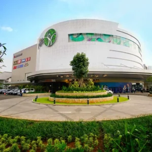 Tampilan luar pusat perbelanjaan (mal) modern bertingkat dengan bangunan melengkung berwarna putih, area taman bundar di depan pintu masuk, dan langit cerah. Gambar ini berada dalam artikel "The Park Sawangan" di website Ecotown.id